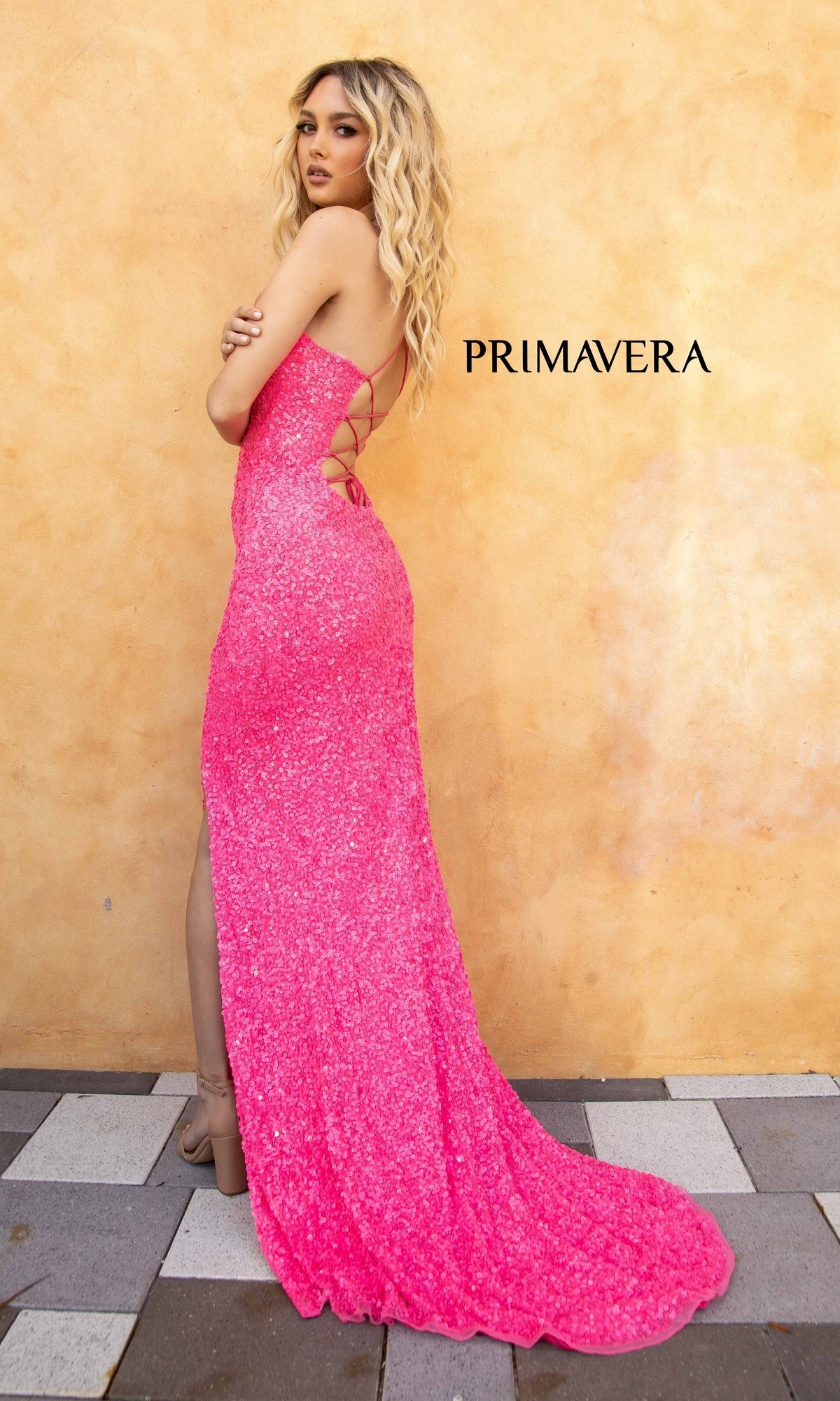 A woman in a vibrant sequin prom dress, the Backless Bright Color Long Sequin Prom Dress 3290B, poses by a textured beige wall featuring